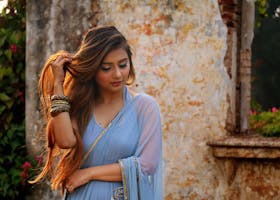 A woman in a blue saree poses gracefully outdoors by a rustic wall.